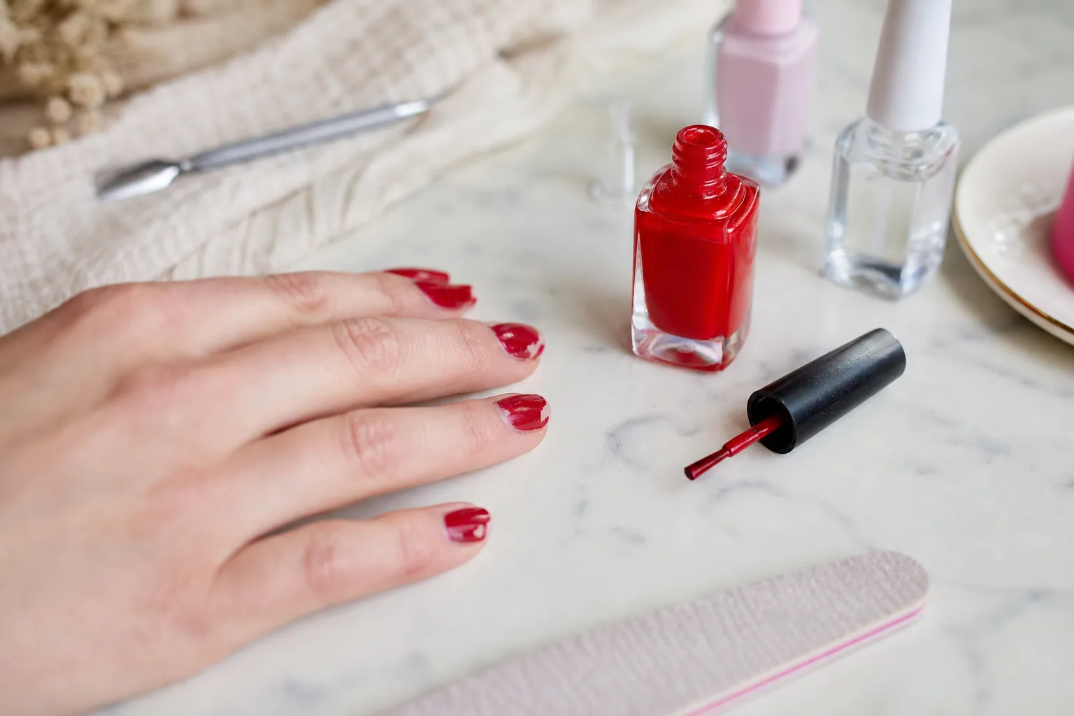 Flacon de vernis à ongles rouge ouvert posé à côté d'une main aux ongles fraîchement vernis, lumière douce